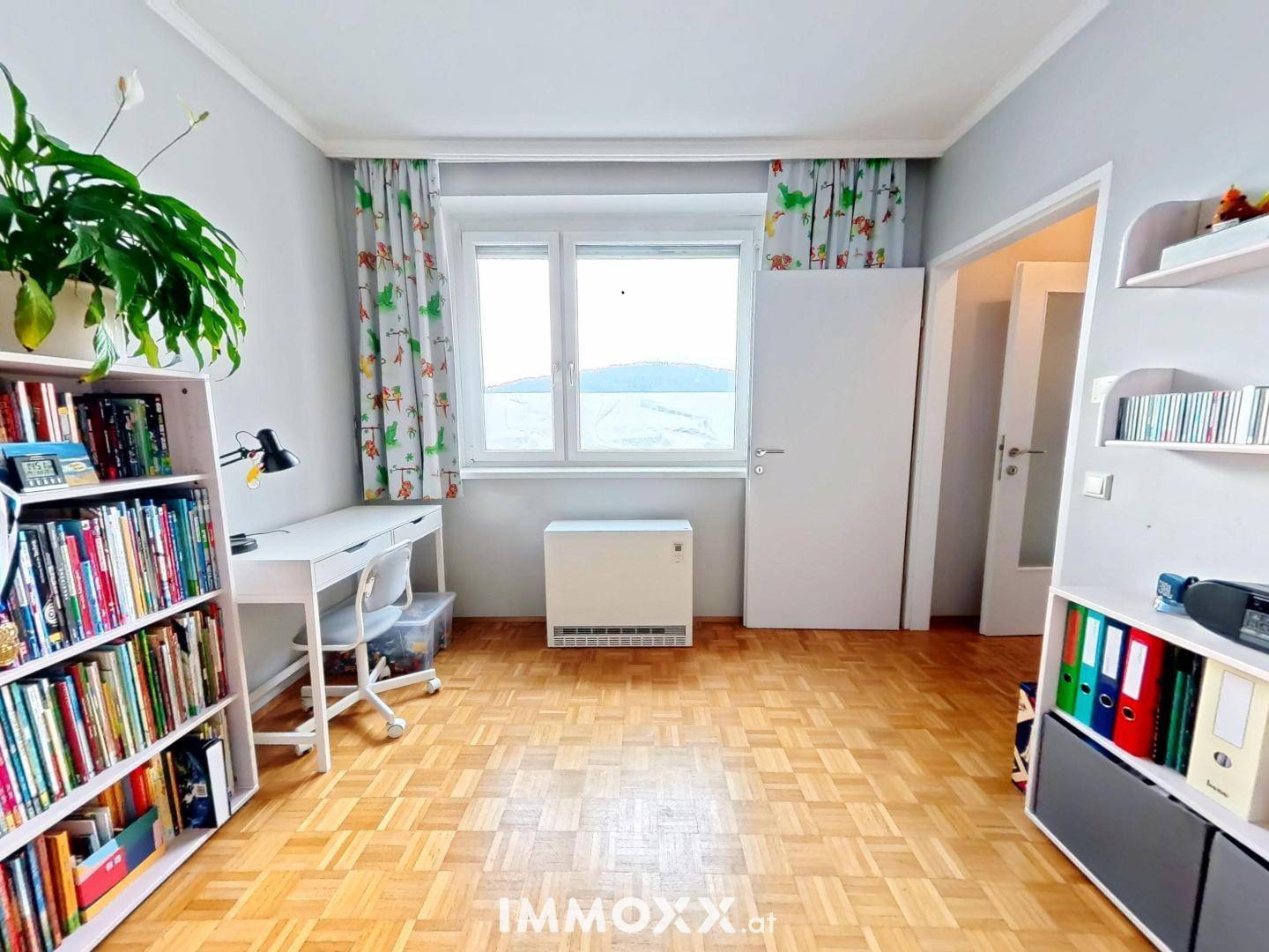 Helles Zimmer mit Parkettboden, Schreibtisch, Bücherregalen und einem Fenster mit Ausblick.