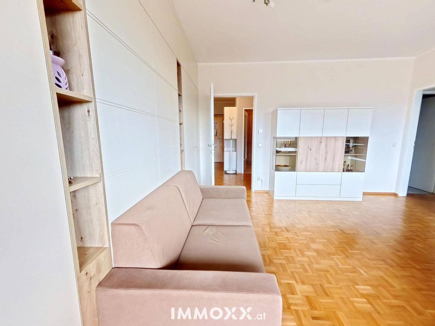Geräumiges Wohnzimmer mit Sofa, Einbauschrank und hellem Parkettboden, offen zum Flur.