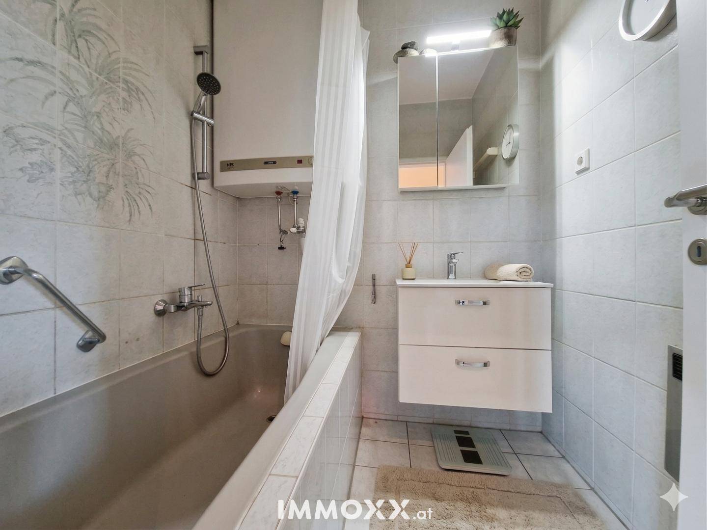 Badezimmer mit Badewanne, Duschvorhang, Haltegriff und Waschbecken auf hell gefliestem Boden.