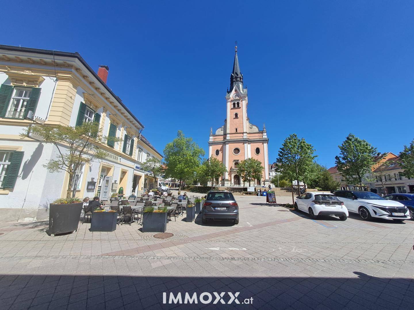 Ein sonniger Stadtplatz mit einer historischen Kirche, Parkplätzen und Außengastronomie.