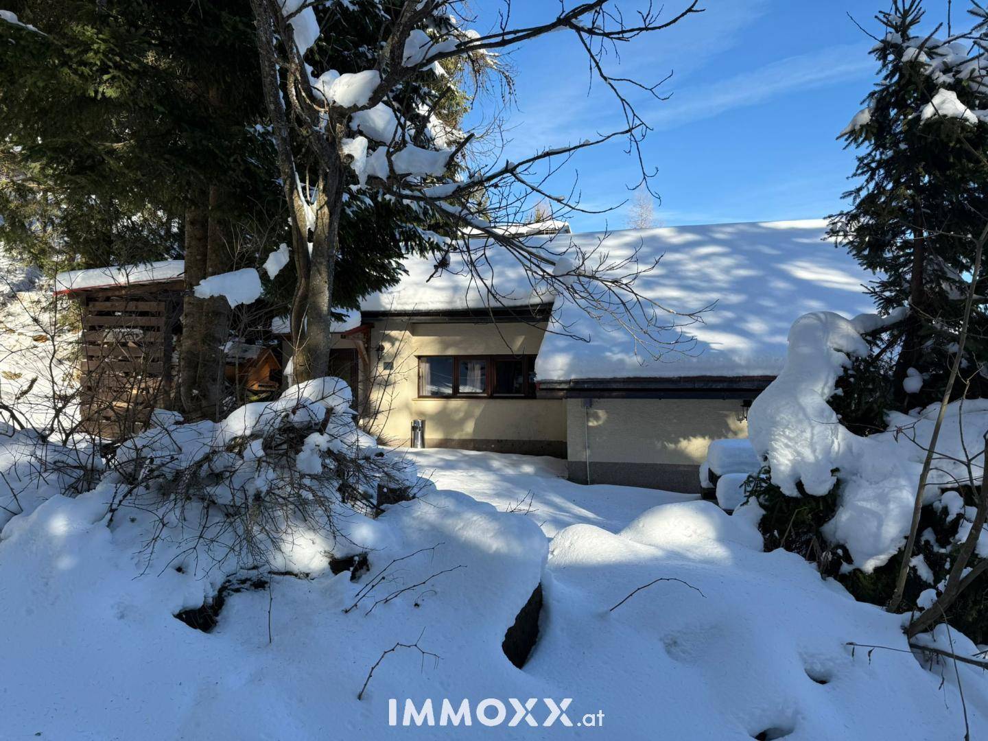 Das Haus ist von tiefem Schnee und hohen Bäumen umgeben, was eine idyllische Winterlandschaft schafft.