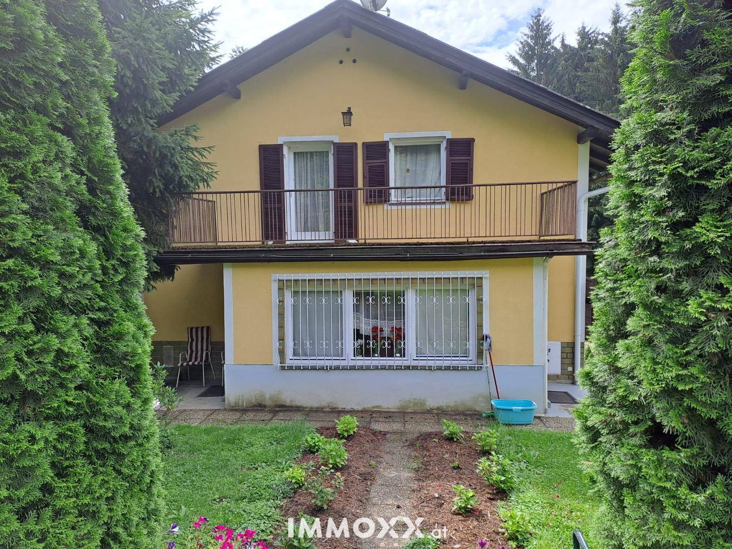 Vorderansicht des gelben Hauses mit Balkon, Fensterläden und einem kleinen Blumenbeet im Vorgarten.