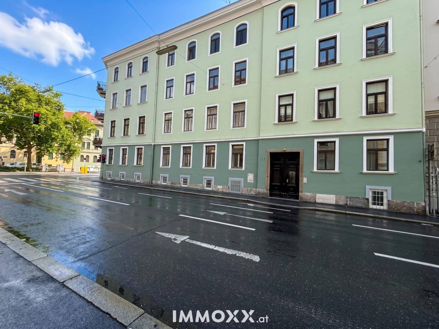 Mehrstöckiges Gebäude mit grüner Fassade und vielen Fenstern, gesehen von einer nassen Straße.