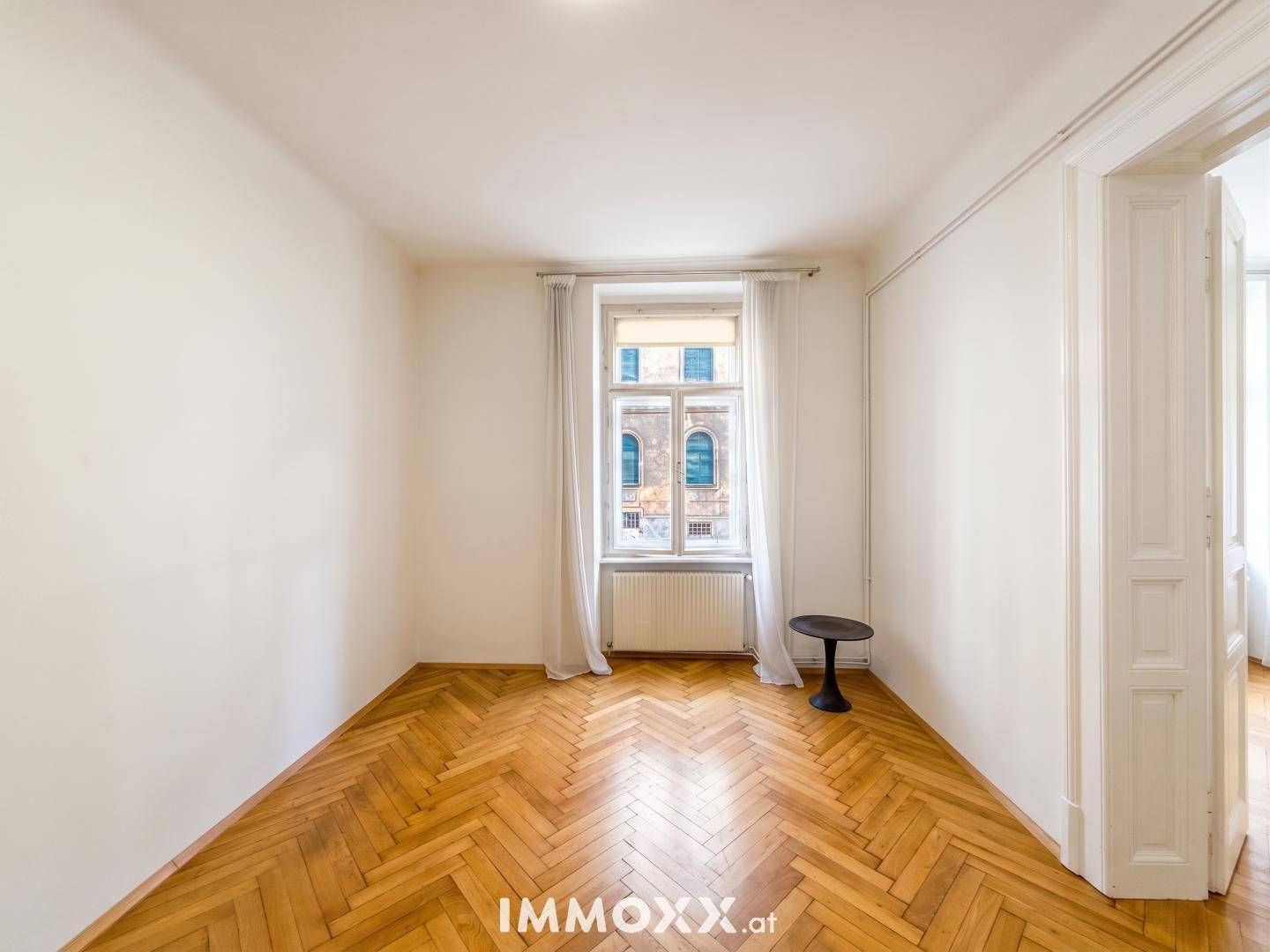 Helles Zimmer mit Fischgrätparkett, großen Fenstern und einem Heizkörper unter dem Fenster.