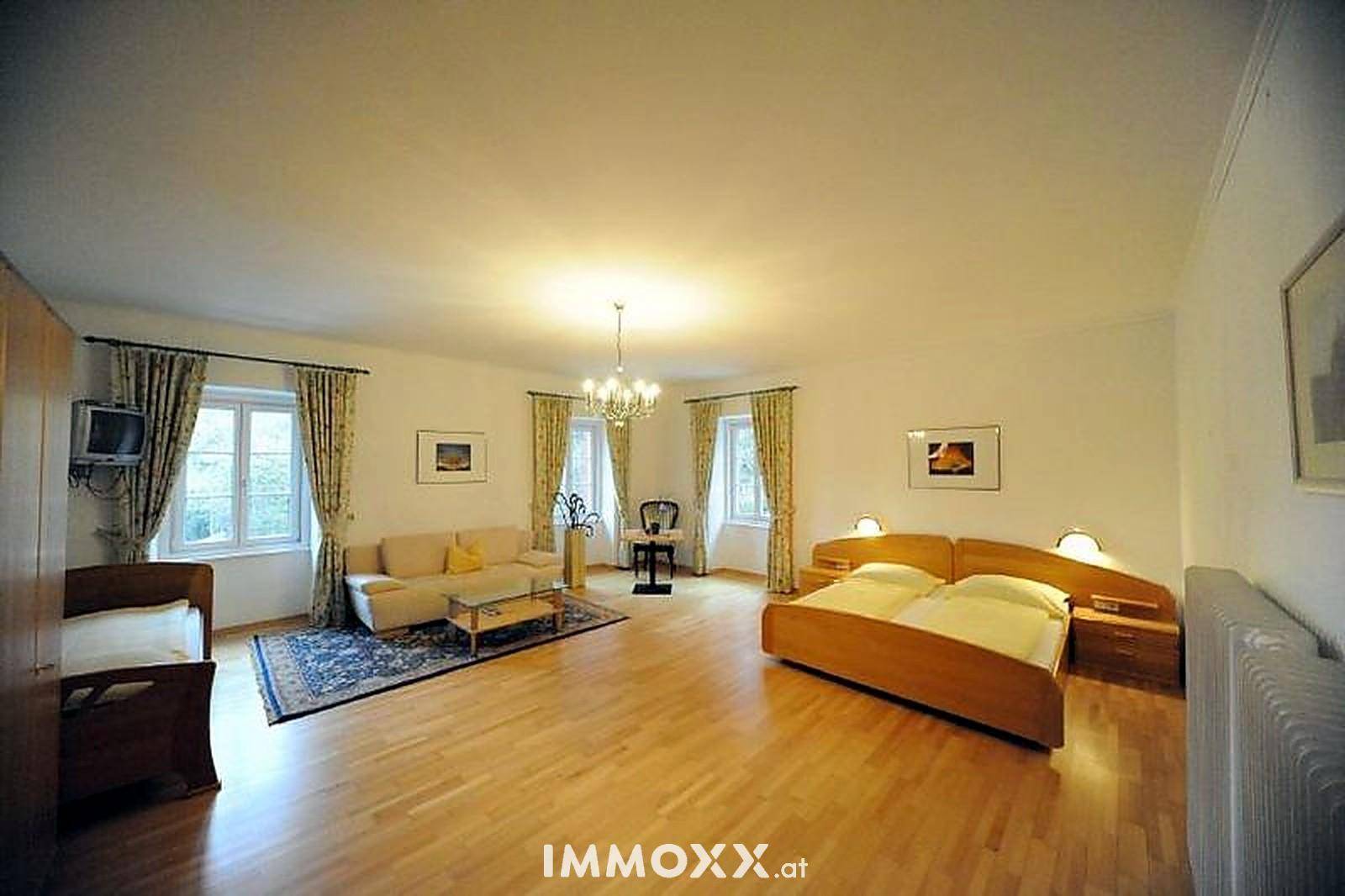 Geräumiges Schlafzimmer mit Doppelbett, Sitzbereich und Holzboden, hell beleuchtet durch mehrere Fenster.