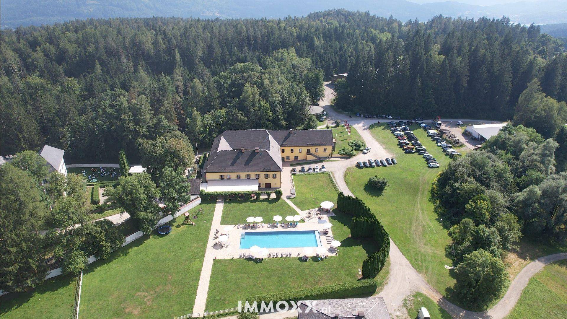 Überblick über das Hauptgebäude, den Außenpoolbereich und den Parkplatz, umgeben von Wald.