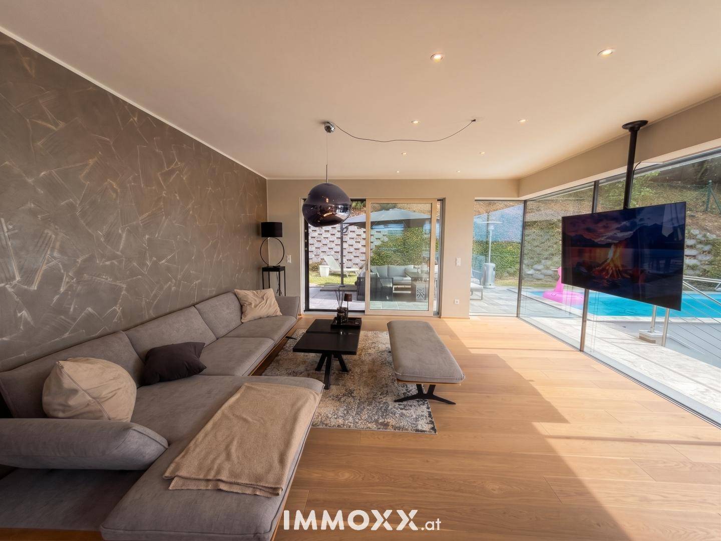 Helles Wohnzimmer mit großem Sofa, Wandfernseher und raumhohen Fenstern zum Pool.