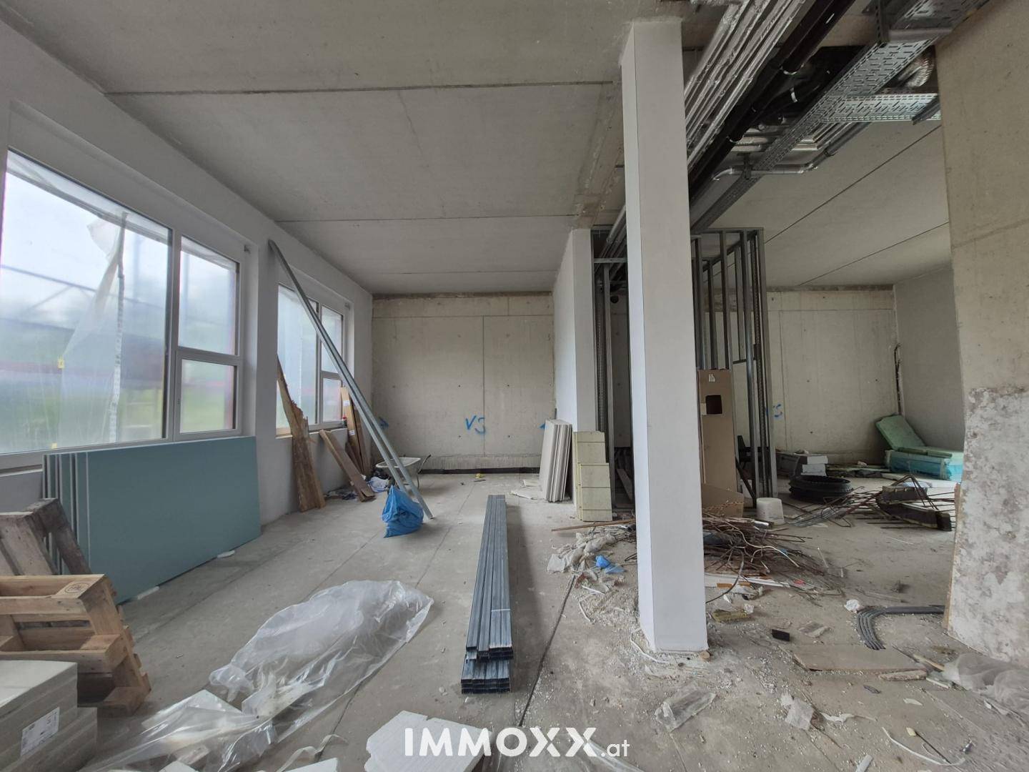 Großer Innenraum im Bau mit vielen Fenstern, Betonstützen und Installationsmaterialien auf dem Boden verteilt.