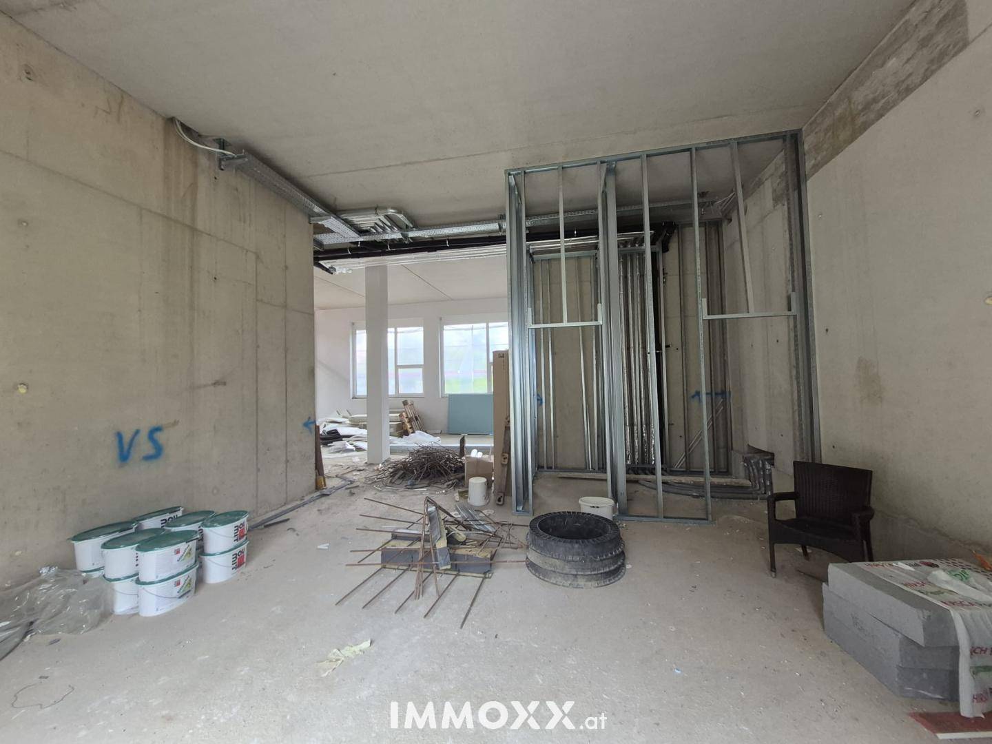 Innenraum im Rohbauzustand mit sichtbaren Betonwänden, Metallständerwerk und Baumaterialien auf dem Boden.