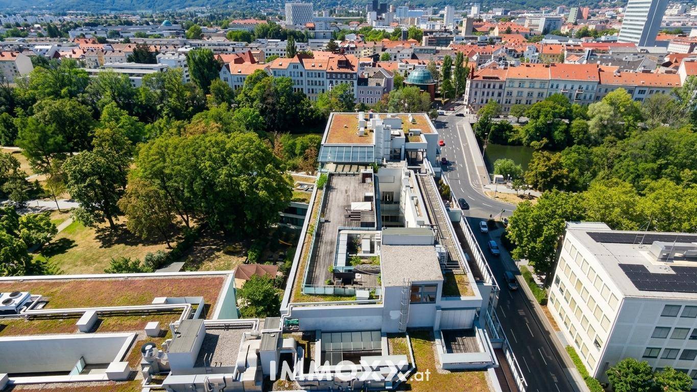 Luftaufnahme eines Gebäudekomplexes mit begrünten Dächern, Bäumen und einer Brücke über den Fluss.