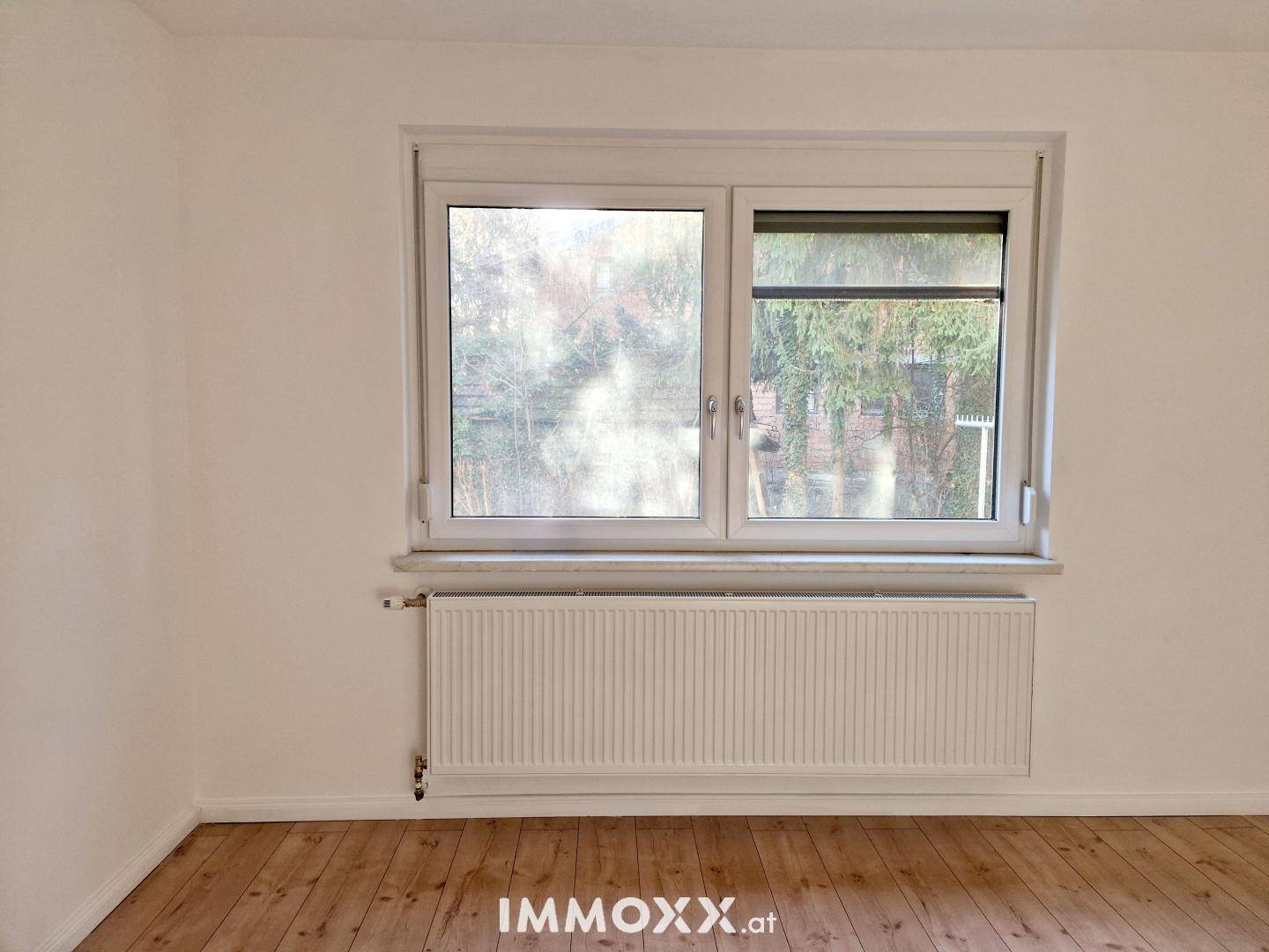 Helles Zimmer mit großem Fenster, Heizkörper und Holzboden, bietet viel Tageslicht.
