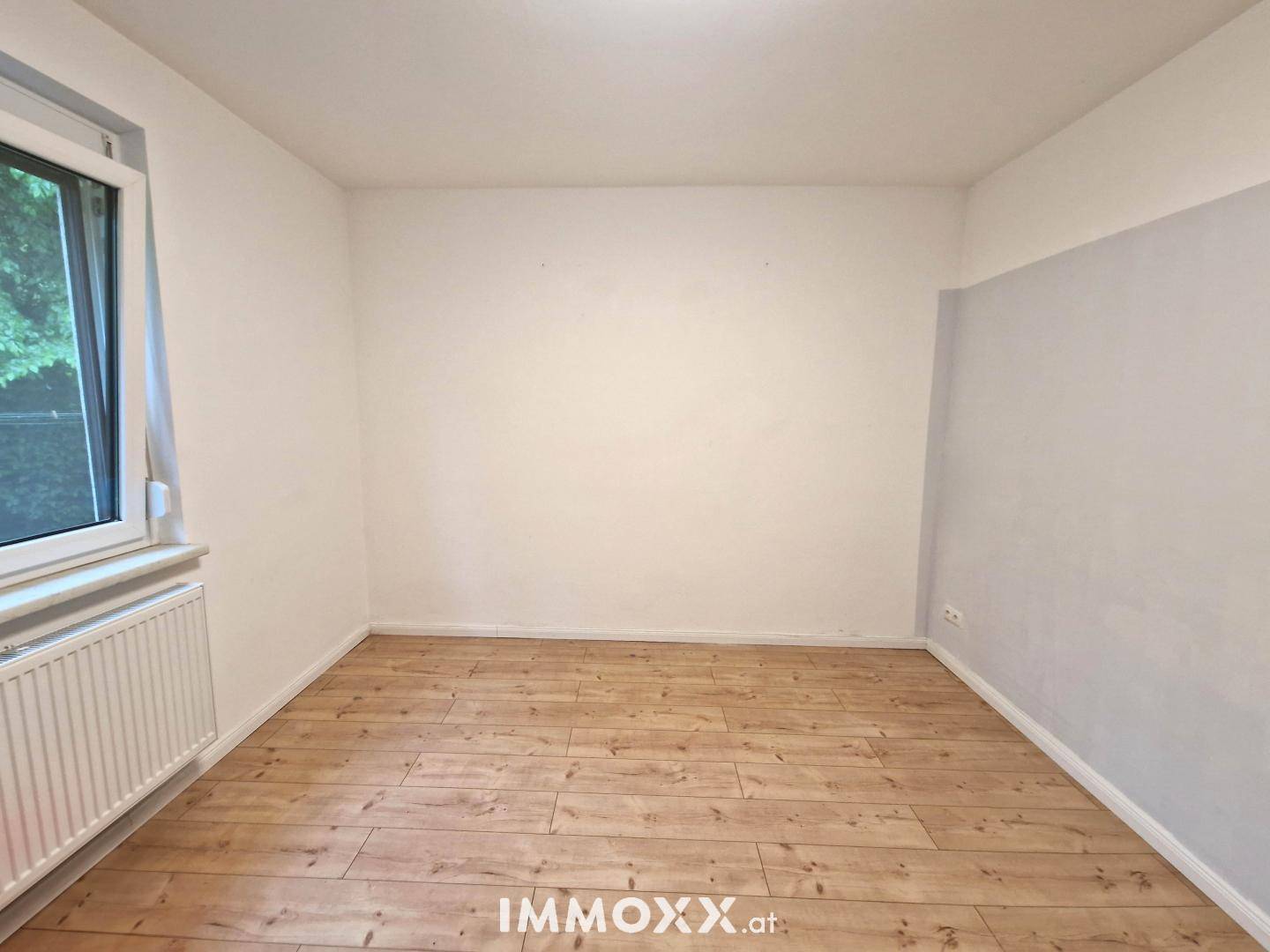 Helles, leeres Zimmer mit Holzboden, weißen Wänden und einem Fenster mit Heizkörper.