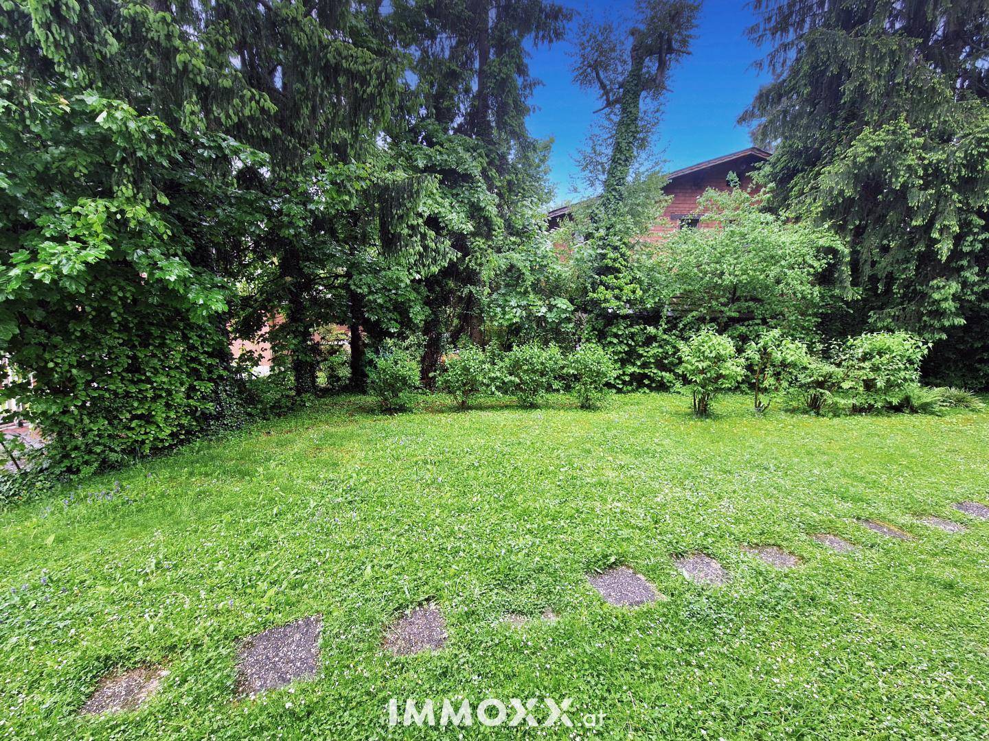Gepflegter Gartenbereich mit Rasenfläche, Trittsteinen und altem Baumbestand im Hintergrund.