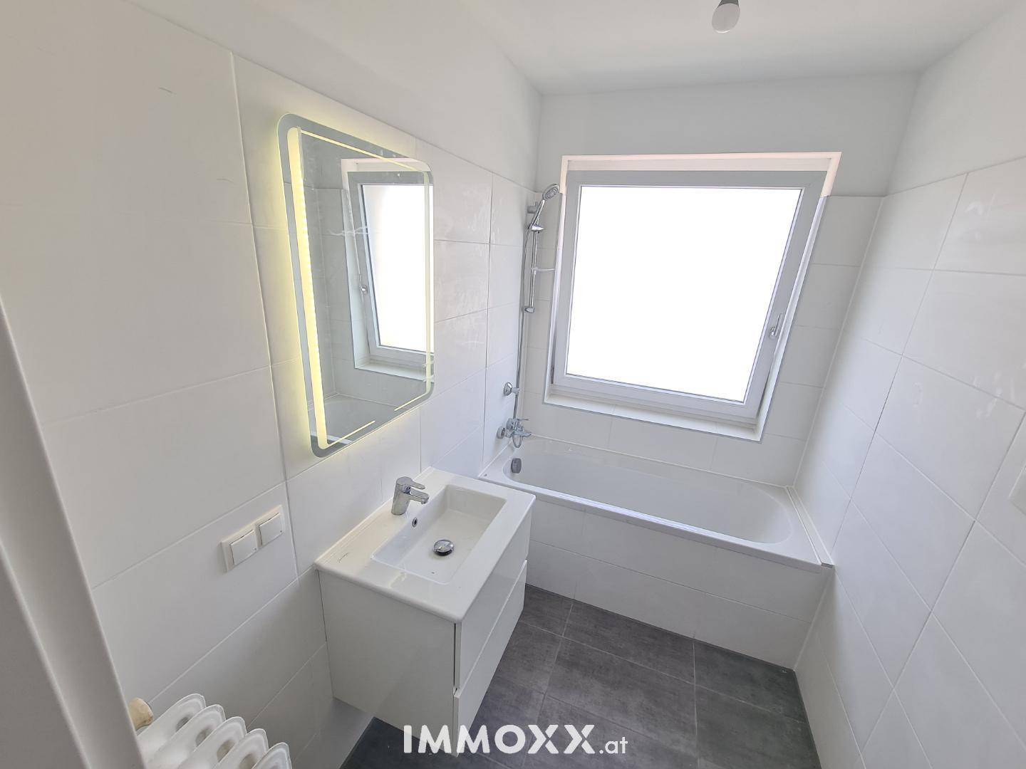 Helles Badezimmer mit Badewanne, Waschbecken, beleuchtetem Spiegel und einem Fenster für Tageslicht.