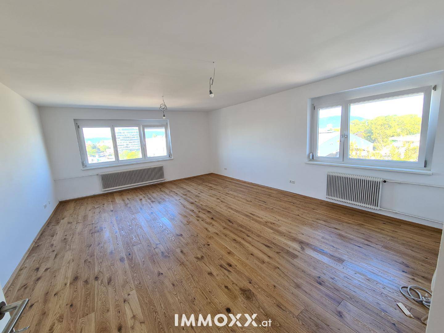 Großes, helles Zimmer mit Holzboden und zwei Fenstern, die viel Tageslicht hereinlassen.