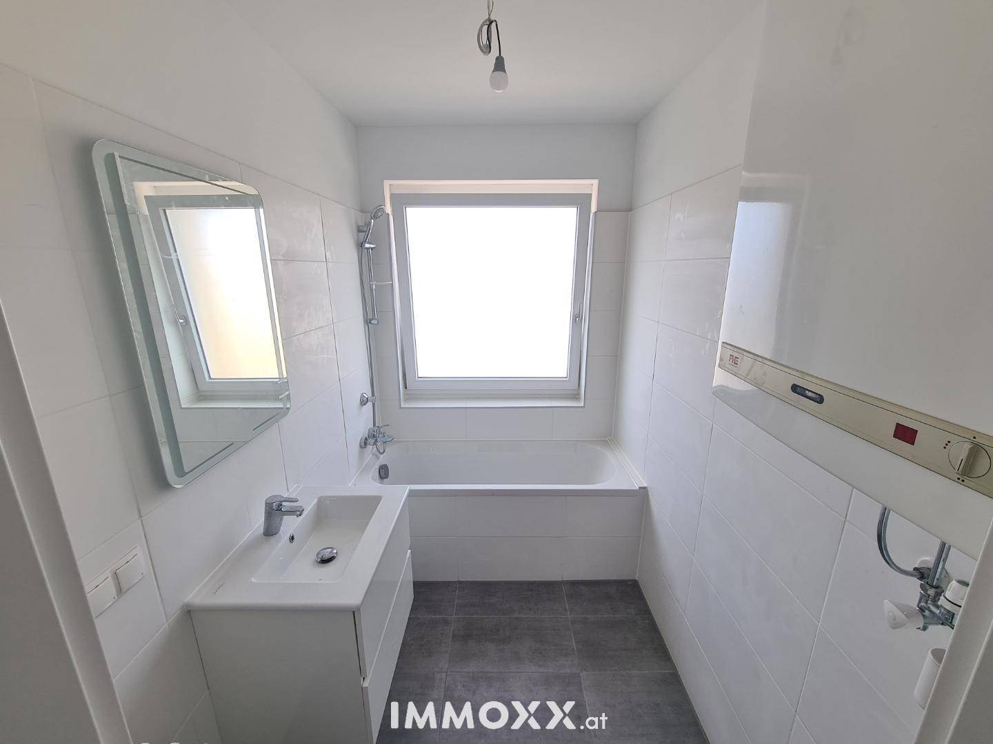 Lichtdurchflutetes Badezimmer mit Badewanne, Waschbecken, Spiegel und Fenster, sowie einem Warmwasserboiler.