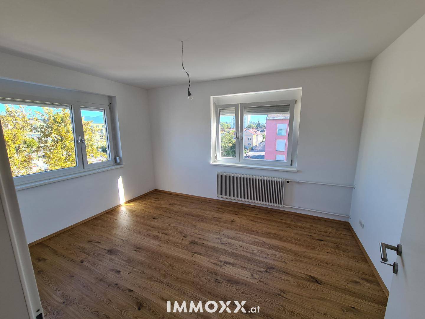Helles Schlafzimmer mit Holzboden und zwei Fenstern, die einen Blick ins Grüne ermöglichen.