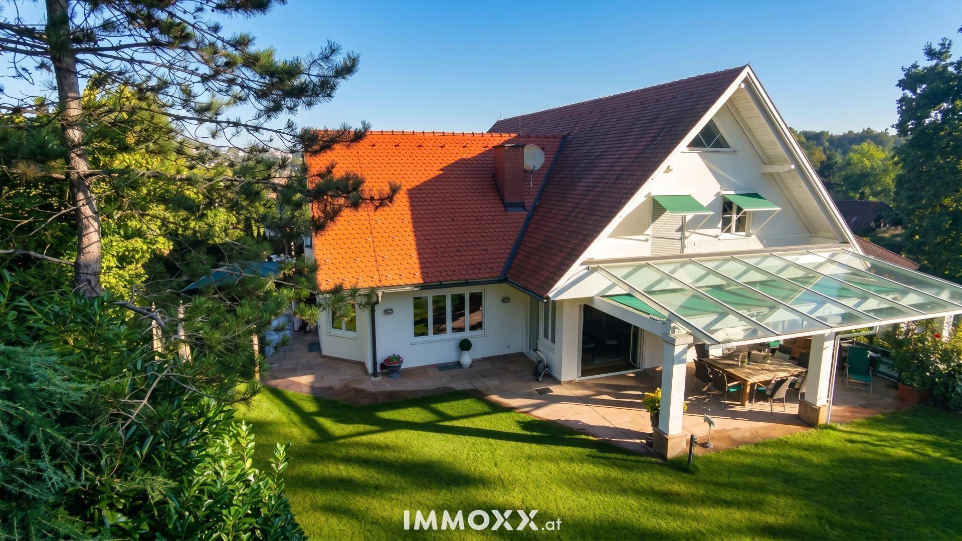 Hausfassade mit überdachter Terrasse und weitläufigem, gepflegtem Rasenbereich im Sonnenschein.
