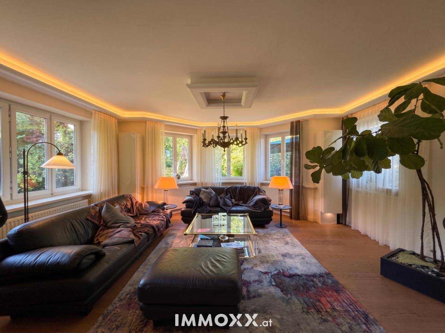 Gemütliches Wohnzimmer mit dunklen Ledersofas, Glastisch und vielen Fenstern für helles Ambiente.