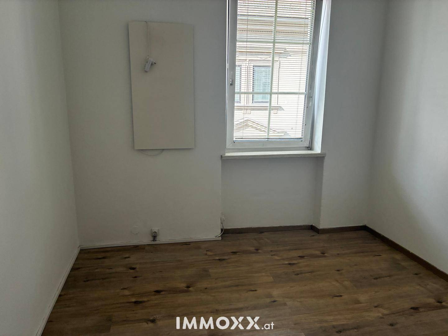 Leerer Raum mit hellen Wänden, Laminatboden und einem Fenster mit Jalousien.