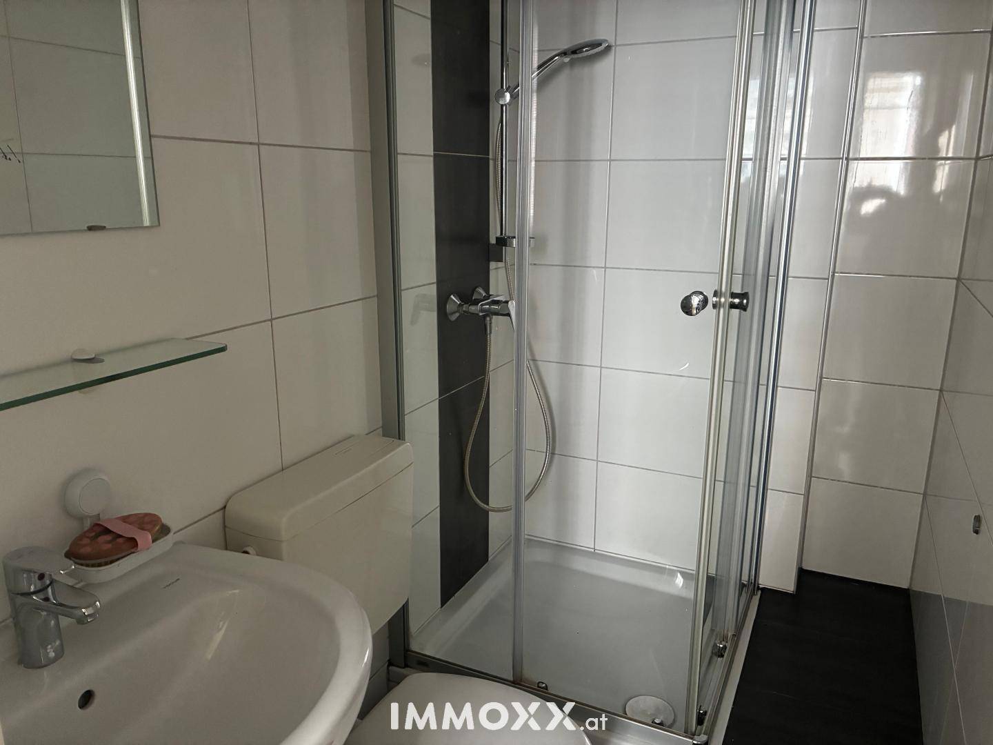 Kleines Badezimmer mit weiß-grauen Fliesen, Waschbecken, Toilette und Duschkabine.