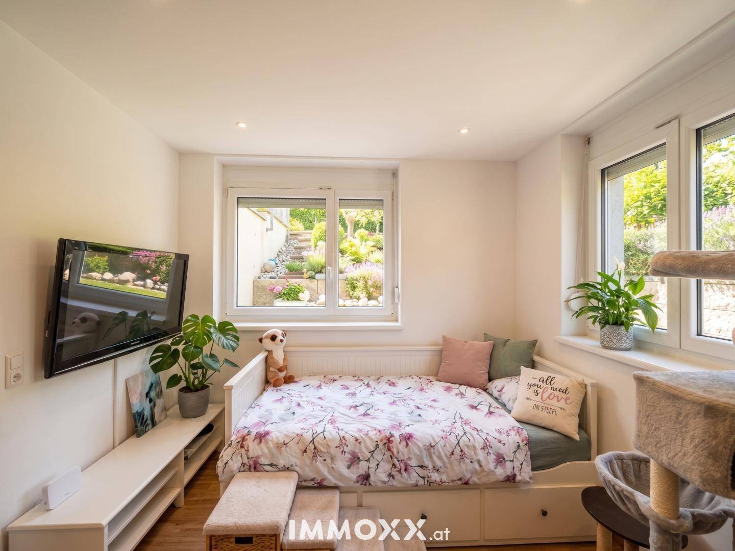 Helles Schlafzimmer mit Bett, Wandfernseher und großen Fenstern mit Blick auf den Garten.