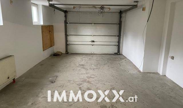 Geräumige Garage mit großem Rolltor, Betonboden, weißen Wänden und kleinen Fenstern.