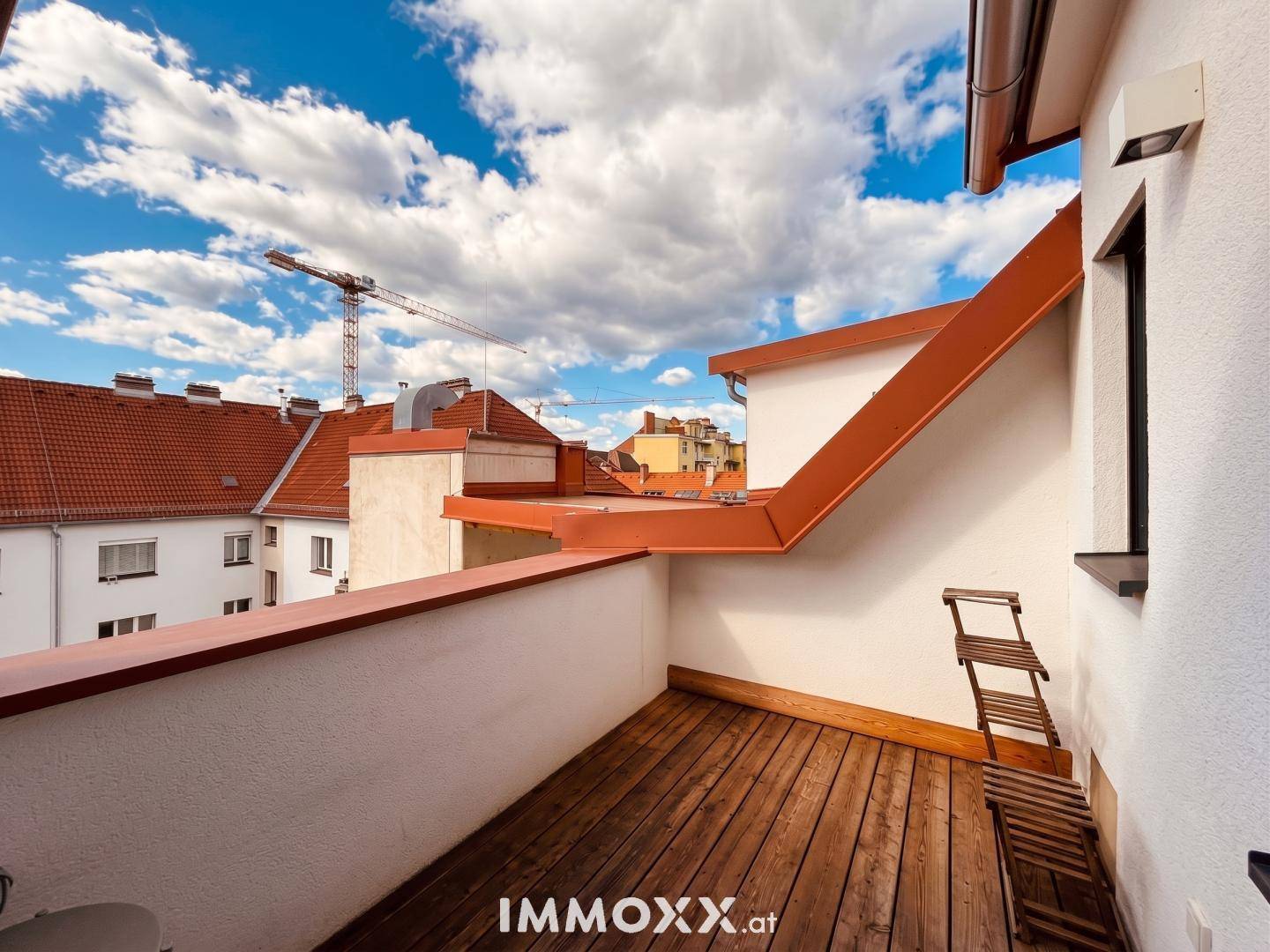 Holzgedeckter Balkon mit Blick auf die umliegenden roten Dächer und einen Baukran am Horizont.