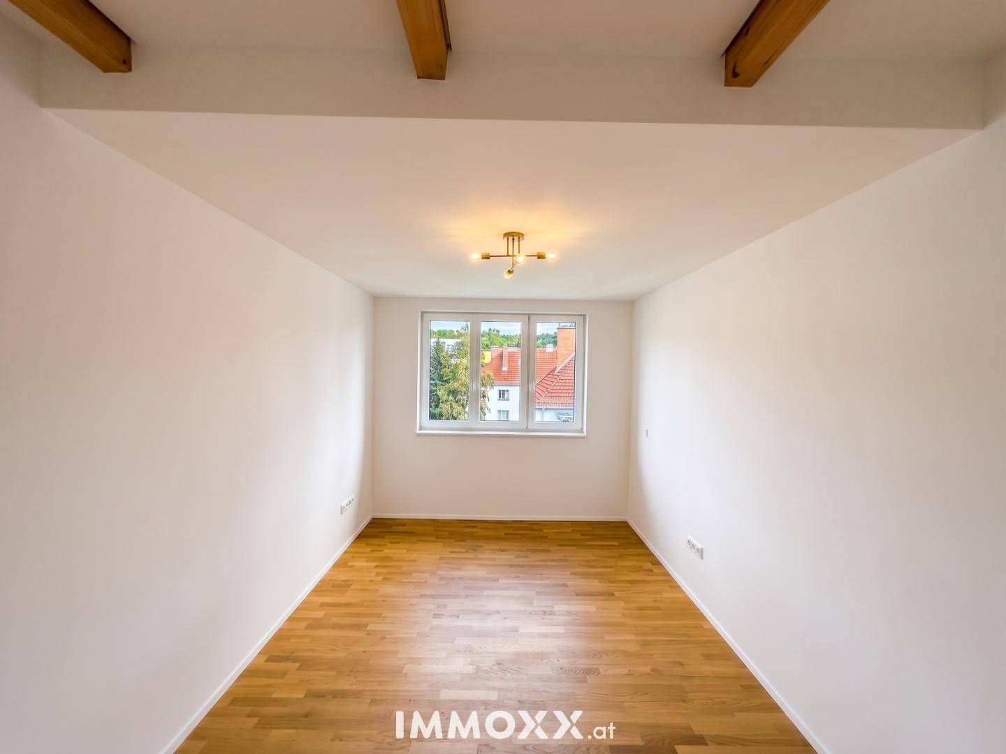 Leeres Zimmer mit Holzboden, Fenster und sichtbaren Holzbalken an der Decke.