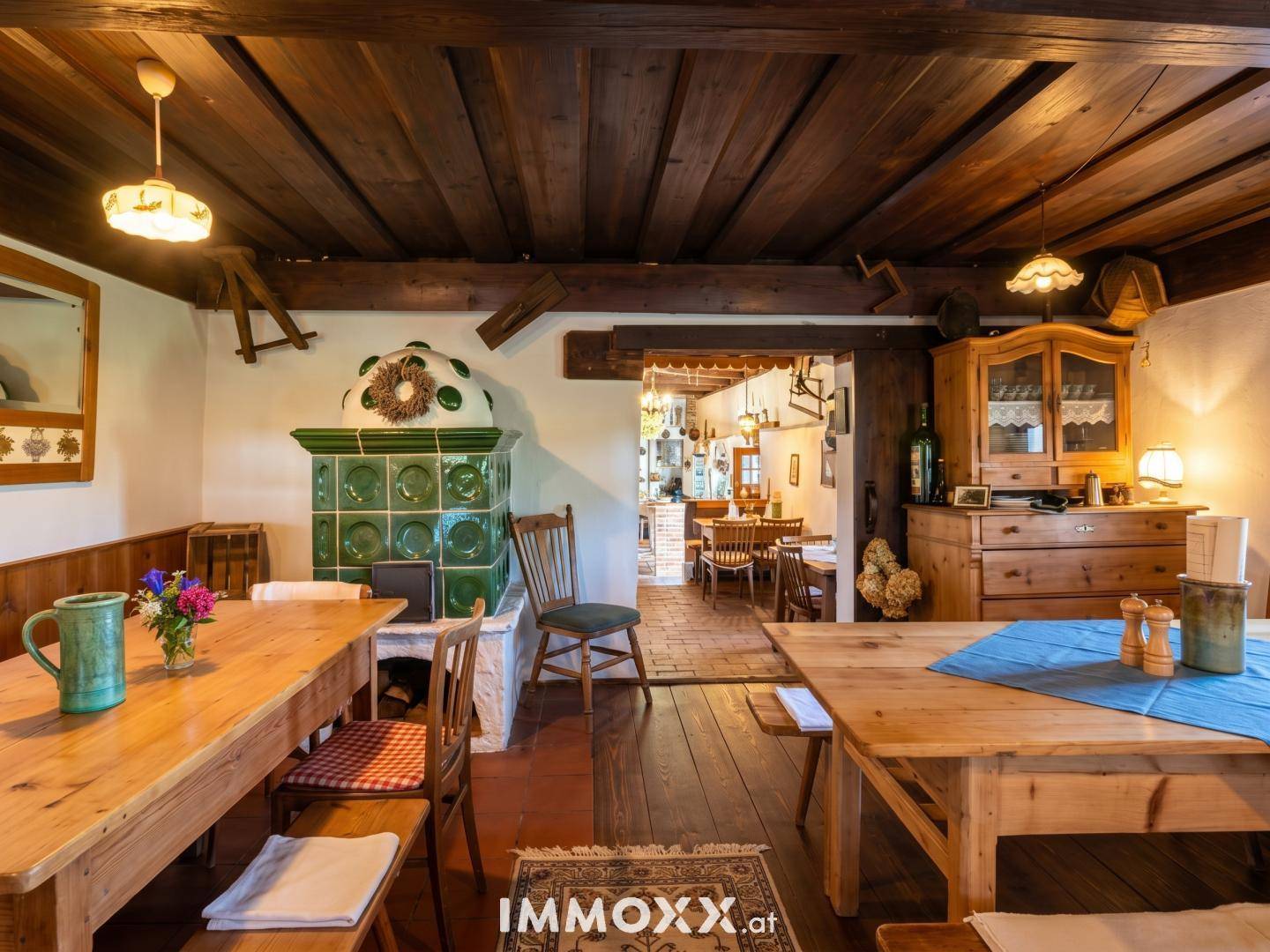 Gemütlicher Speisesaal mit traditionellem Kachelofen, Holztischen und Blick in einen weiteren Raum.