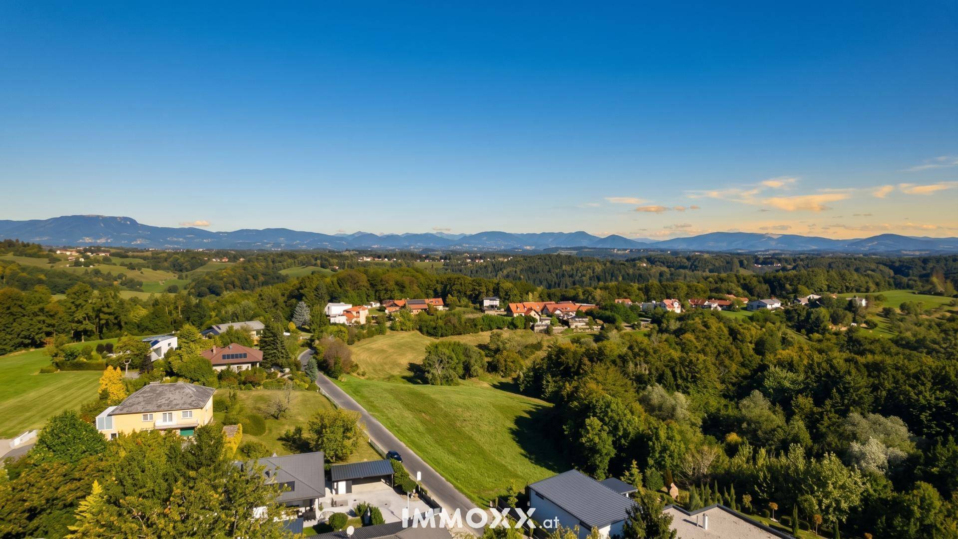 Panoramablick auf eine idyllische Landschaft mit Wohnhäusern, grünen Feldern und einer Bergkette in der Ferne.