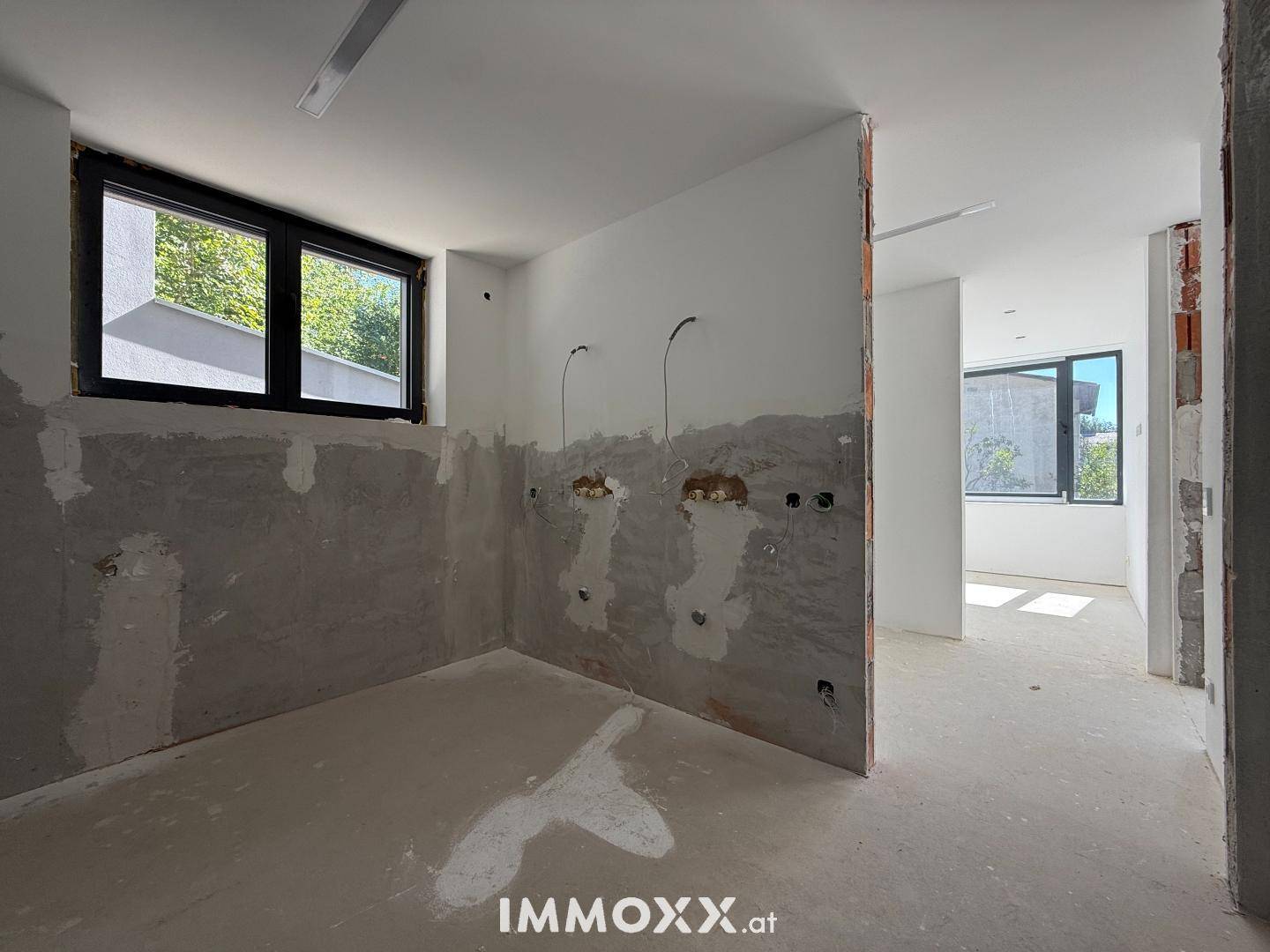 Unfertiger Raum mit Fenster und vorbereiteten Anschlüssen an der Wand, ideal für Bad oder Küche.