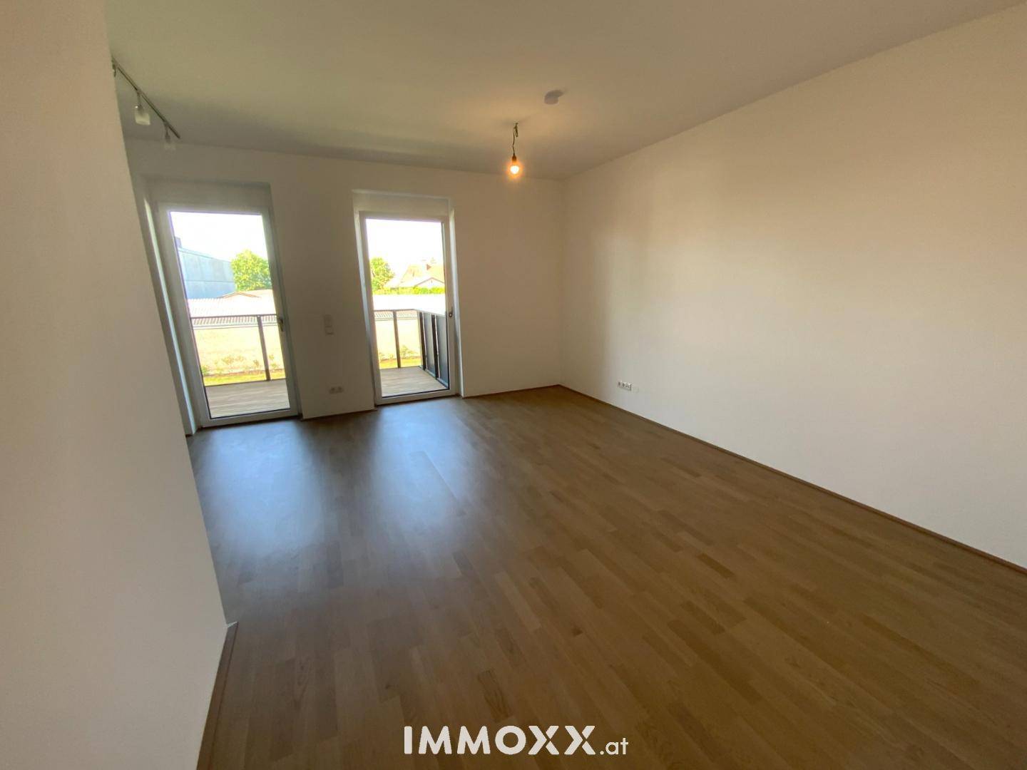 Leerer Wohnraum mit Holzboden und zwei Fenstertüren, die auf einen Balkon führen.