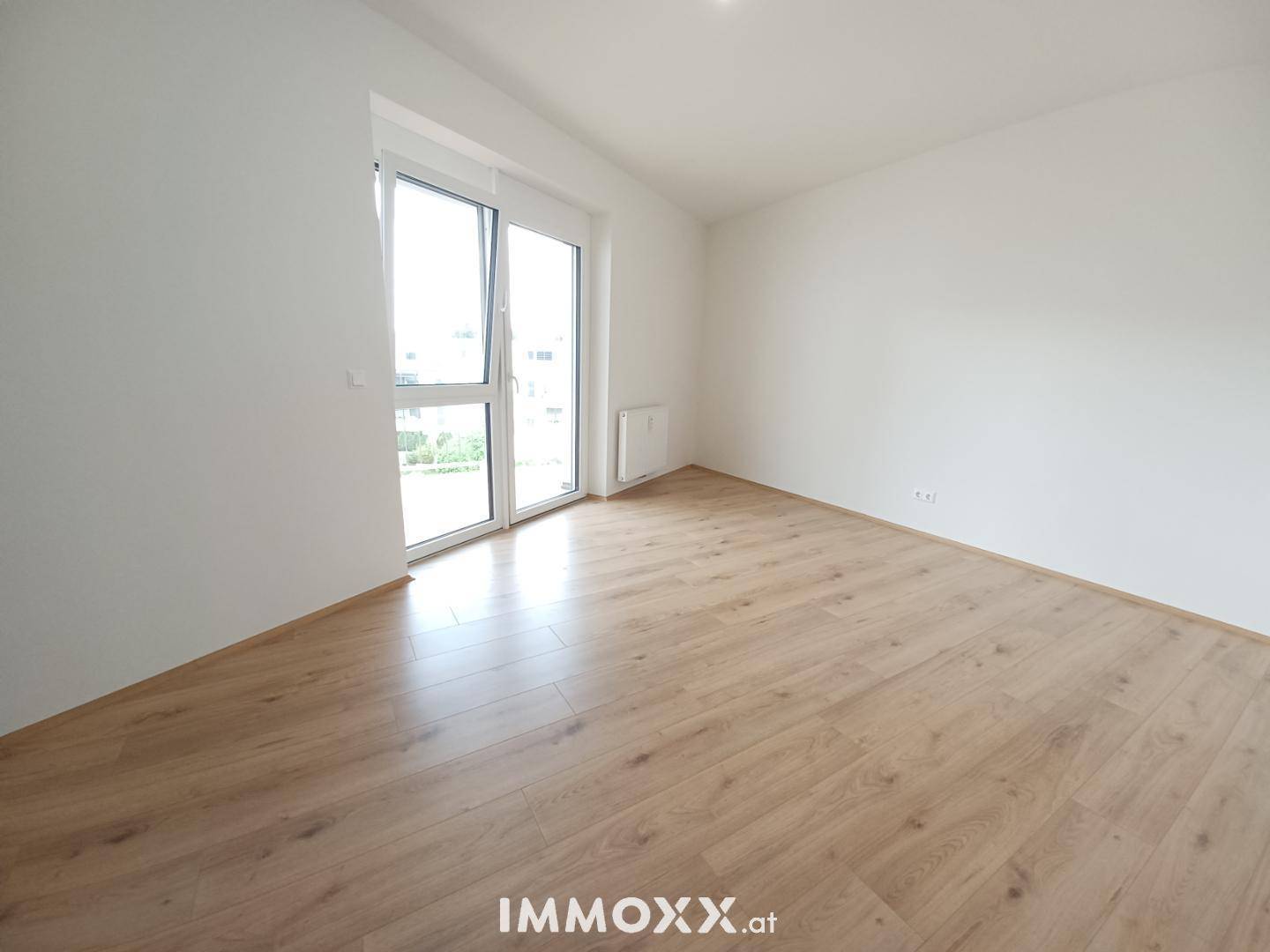 Leeres, helles Zimmer mit Laminatboden und großem Fenster, das viel Tageslicht hereinlässt.