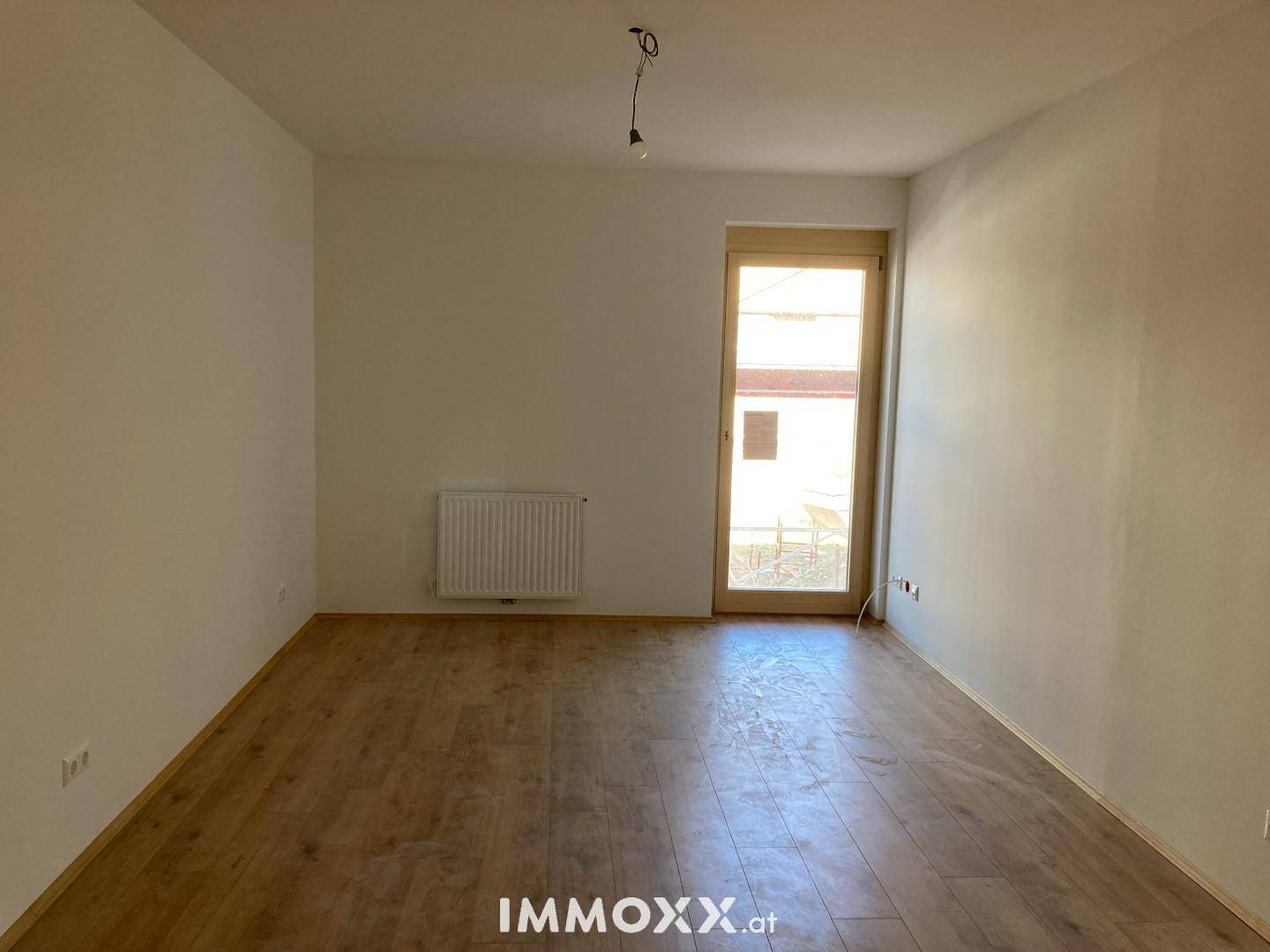 Leerer Raum mit Laminatboden, weißen Wänden und einem Fenster, das viel Tageslicht hereinlässt.