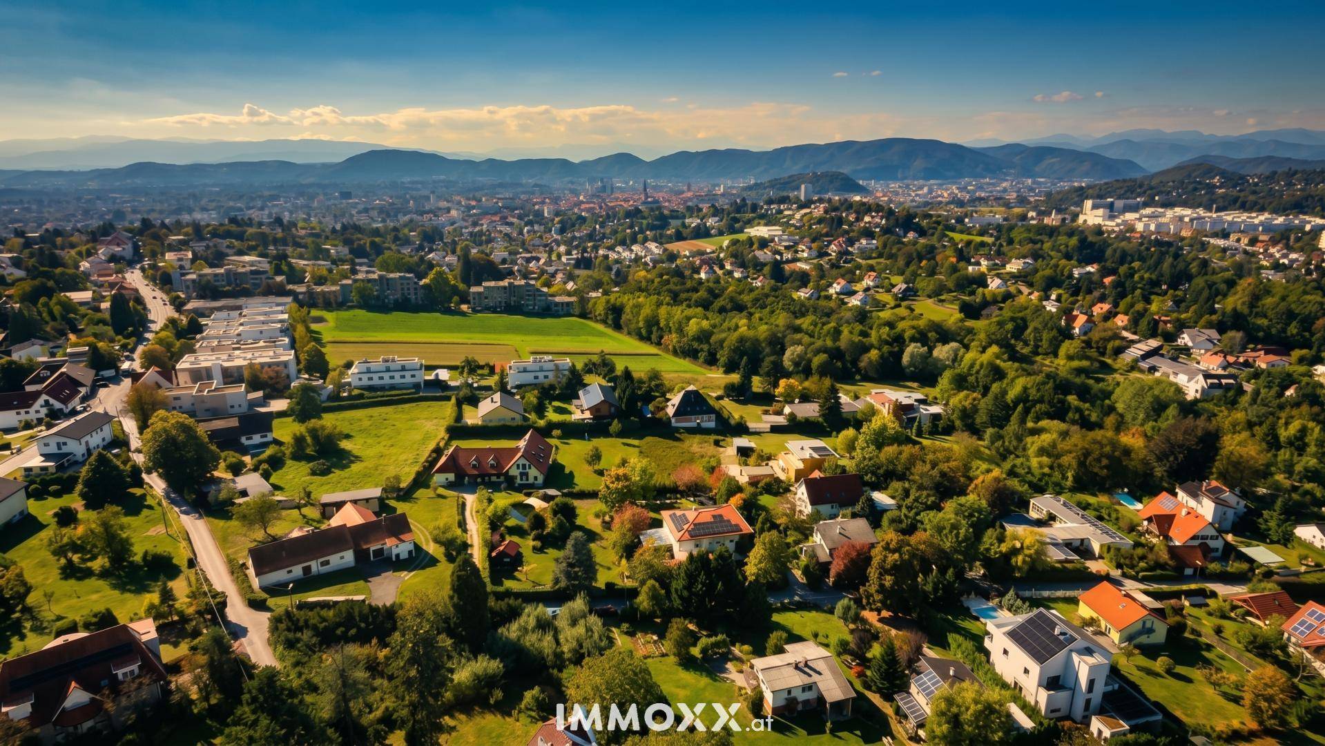 Umfassende Luftaufnahme einer Stadt mit Wohngebieten, weiten Grünflächen und Bergpanorama.