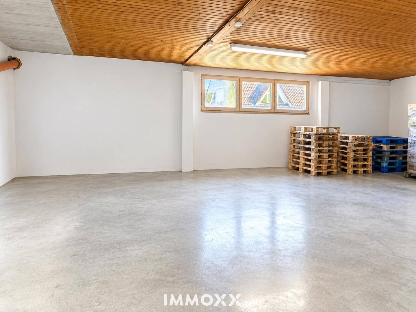 Heller Lagerraum mit Betonboden, weiß gestrichenen Wänden und einer Holzlamellendecke, ausgestattet mit Fenstern.
