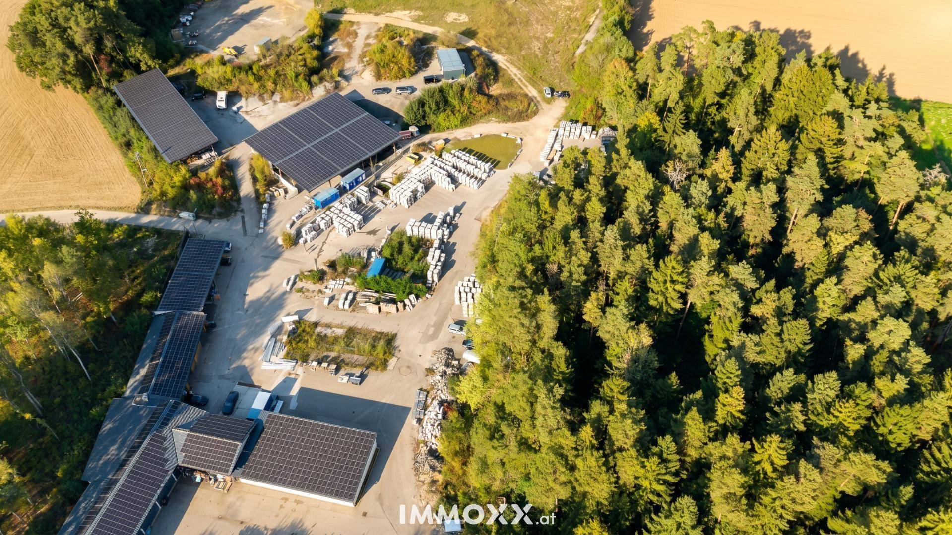 Detaillierte Luftansicht eines Gewerbeareals mit mehreren Photovoltaikdächern und Materiallagerung inmitten grüner Natur.