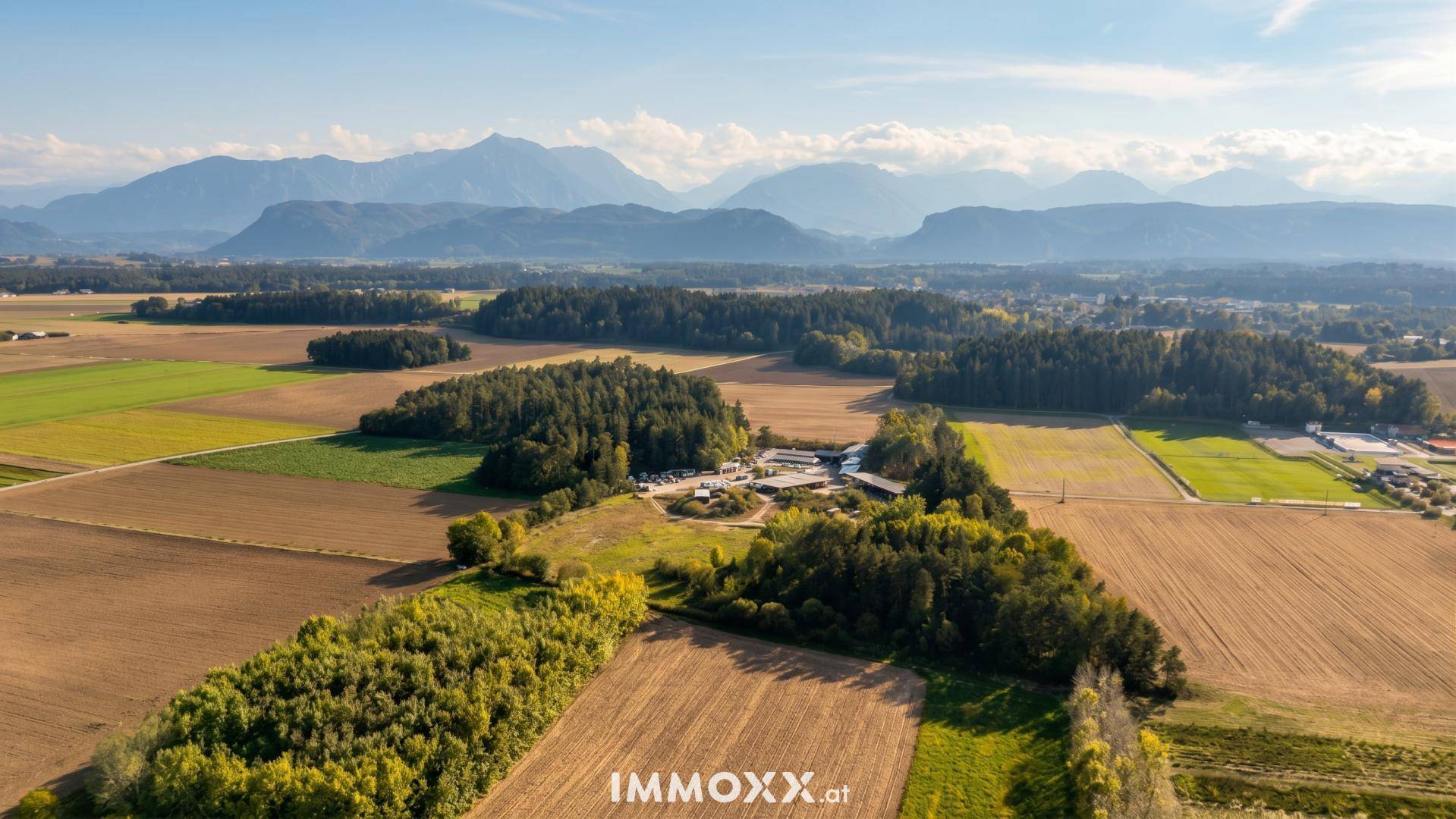Weitläufige Luftaufnahme einer ländlichen Landschaft mit Feldern, Wäldern und majestätischen Bergen im Hintergrund.