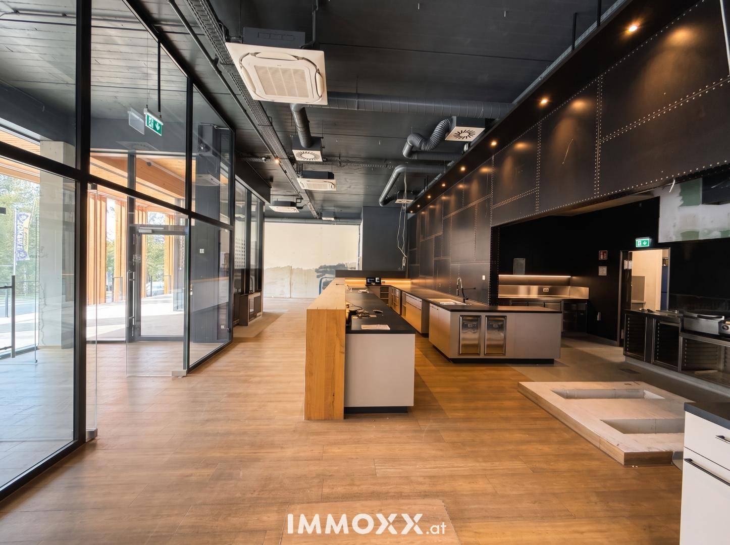 Moderner Küchenbereich eines Restaurants mit Theke, Edelstahloberflächen und großen Glasfronten, viel natürliches Licht.