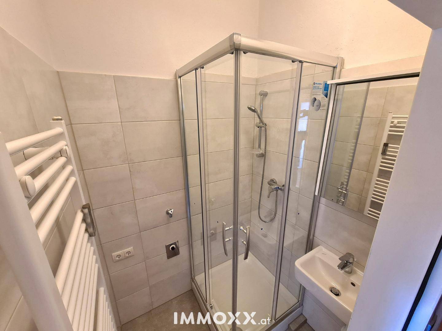 Modernes Badezimmer mit Duschkabine aus Glas, kleinem Waschbecken und beheiztem Handtuchhalter.