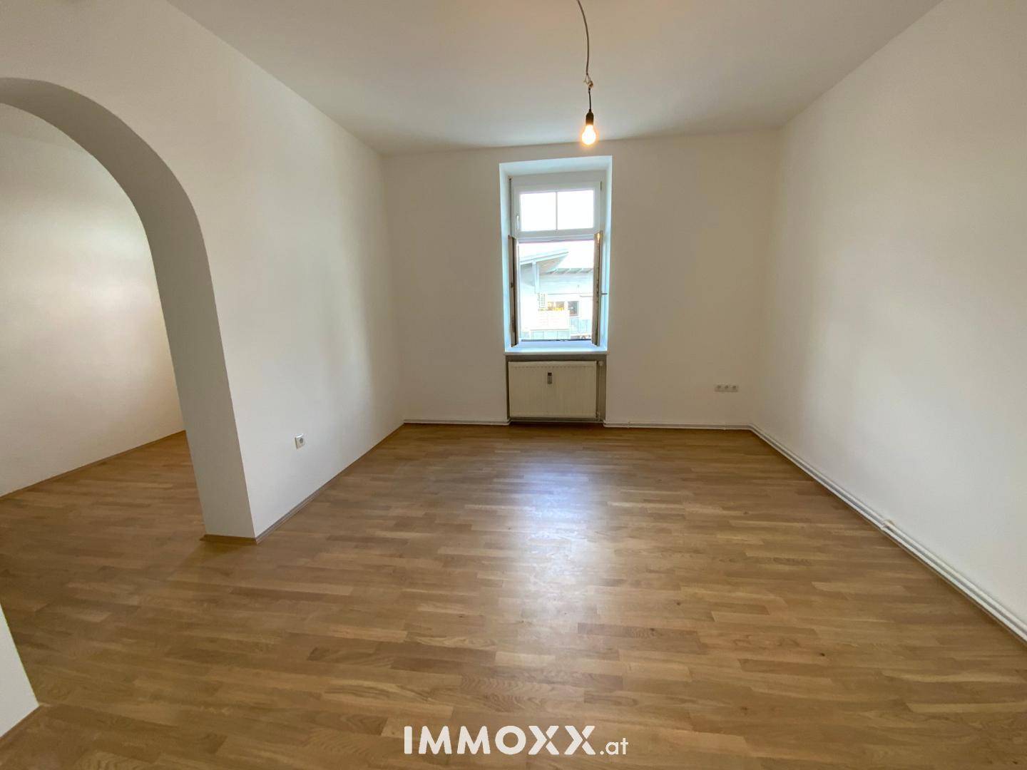 Heller, leerer Wohnraum mit Holzboden, weißen Wänden und einem Fenster mit Blick nach draußen.