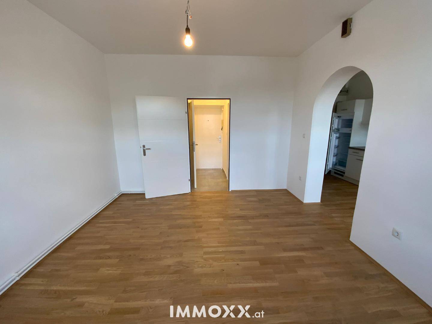 Geräumiger, leerer Wohnraum mit Holzboden, weißen Wänden und Zugang zu Flur und Küche.