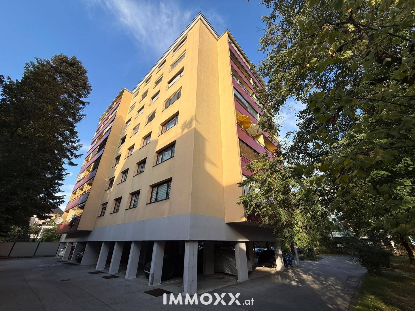 Mehrstöckiges Wohngebäude mit gelber Fassade, Balkonen und überdachten Parkplätzen im Erdgeschoss.