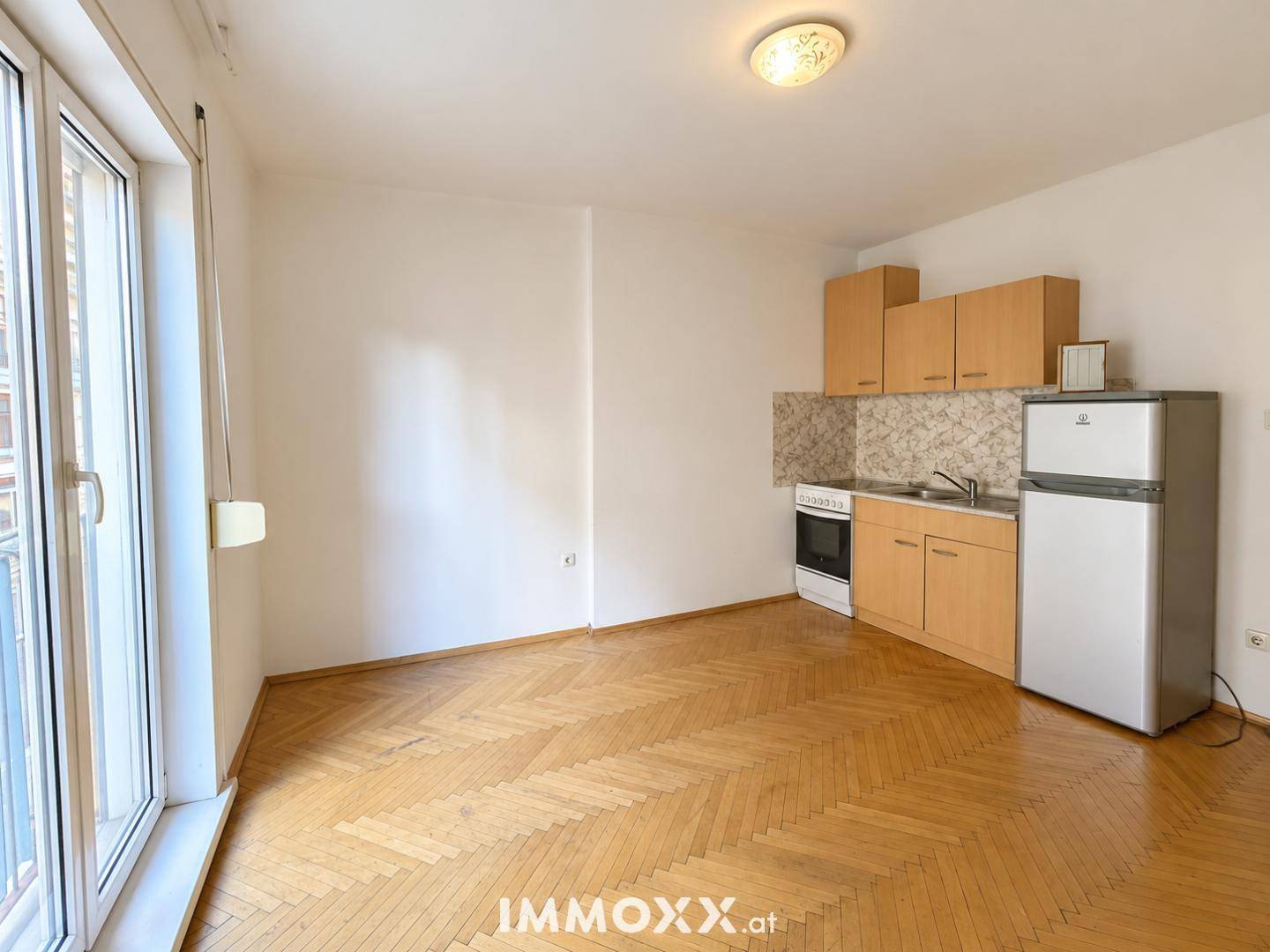 Heller Wohnraum mit integrierter Küchenzeile, Kühlschrank und Fischgrätparkettboden.