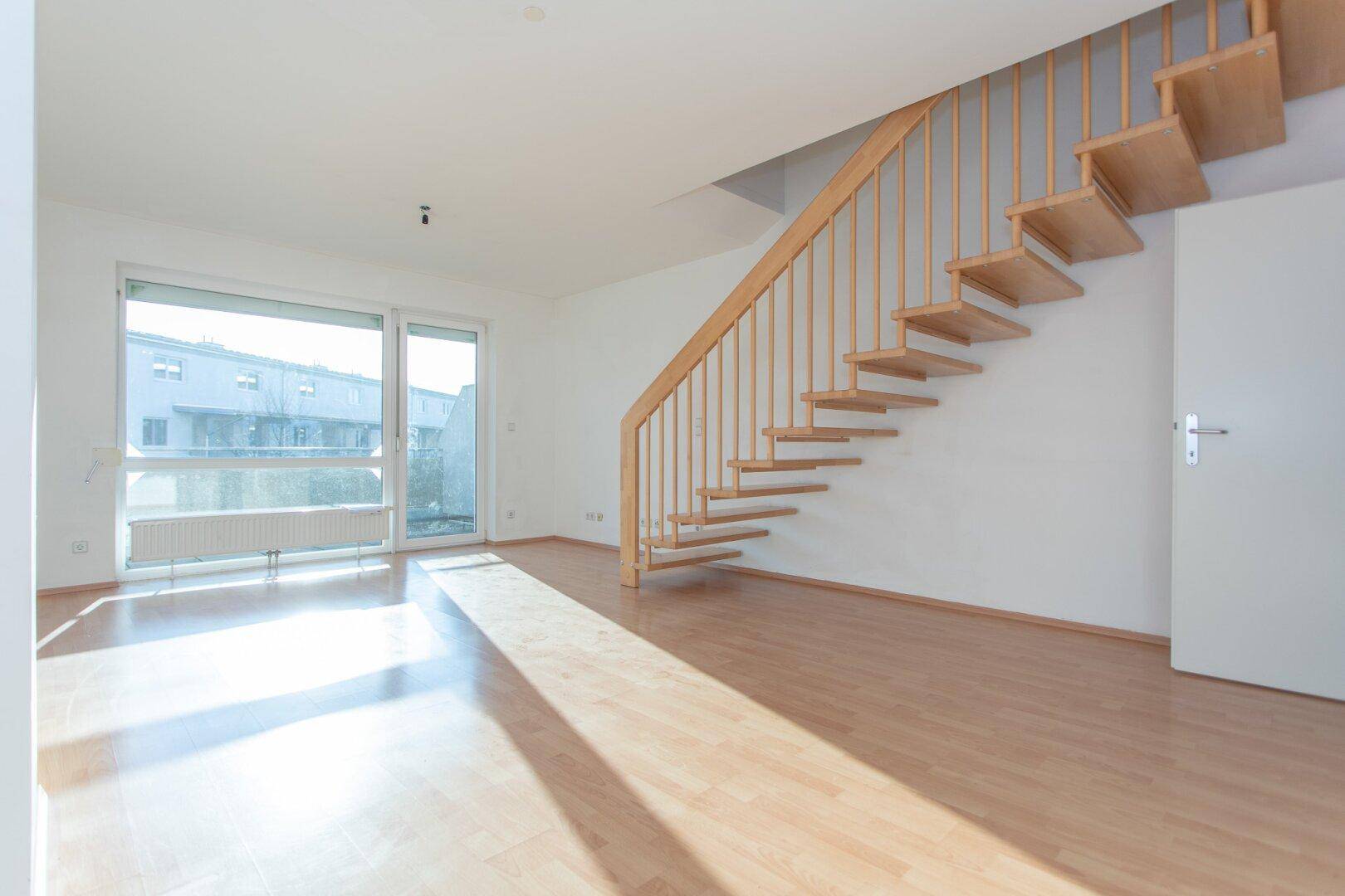 Lichtdurchflutetes Wohnzimmer mit breiter Fensterfront und Holztreppe, bietet viel Platz und Helligkeit.