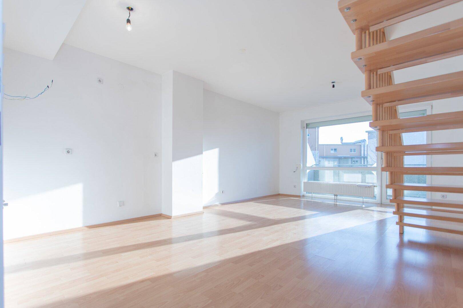 Großzügiges Wohnzimmer mit viel Tageslicht, Holztreppe und Blick nach draußen, bereit zur Einrichtung.