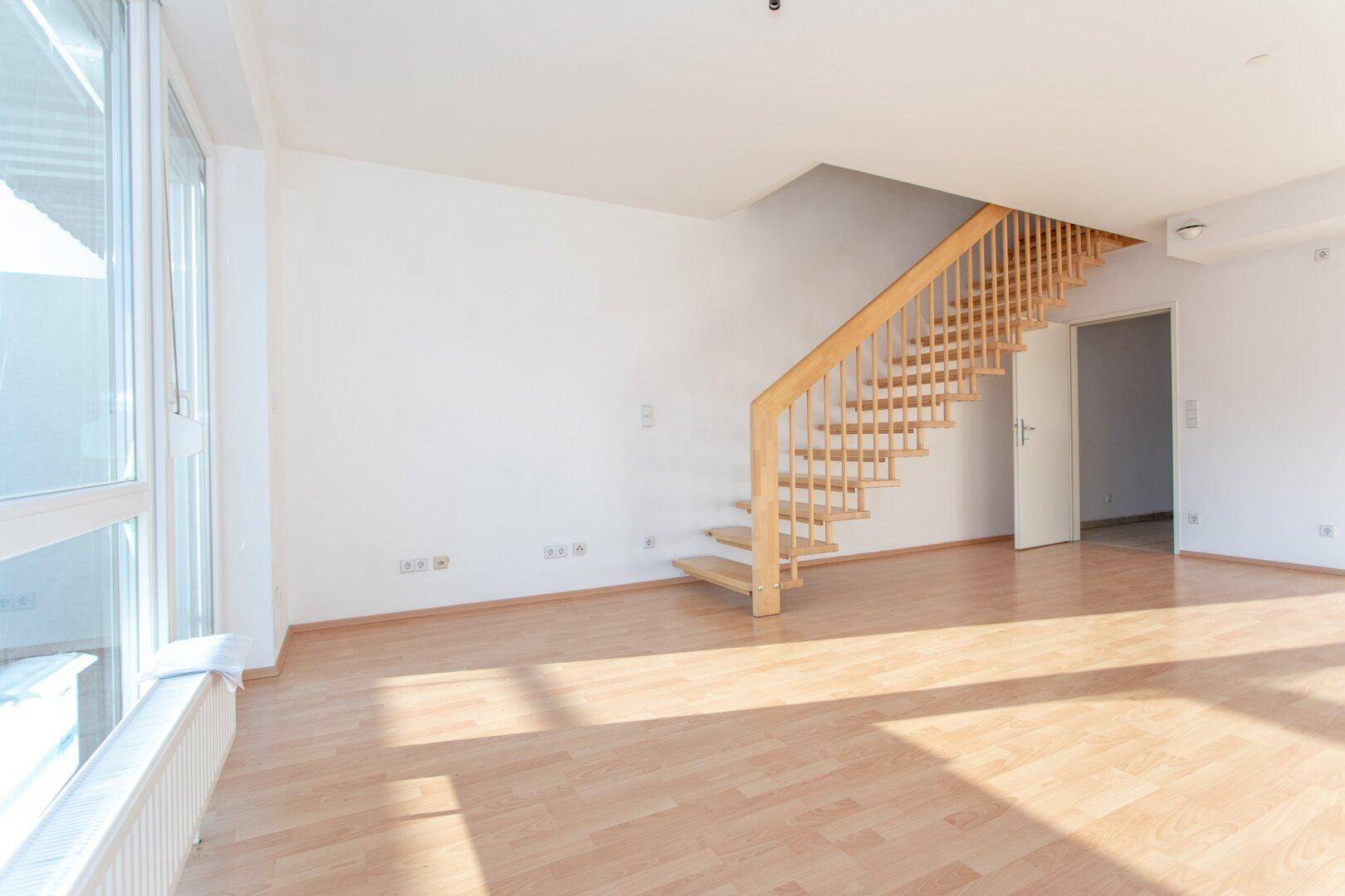 Geräumiges Wohnzimmer mit heller Holztreppe und großen Fenstern, die viel Sonnenlicht hereinlassen.