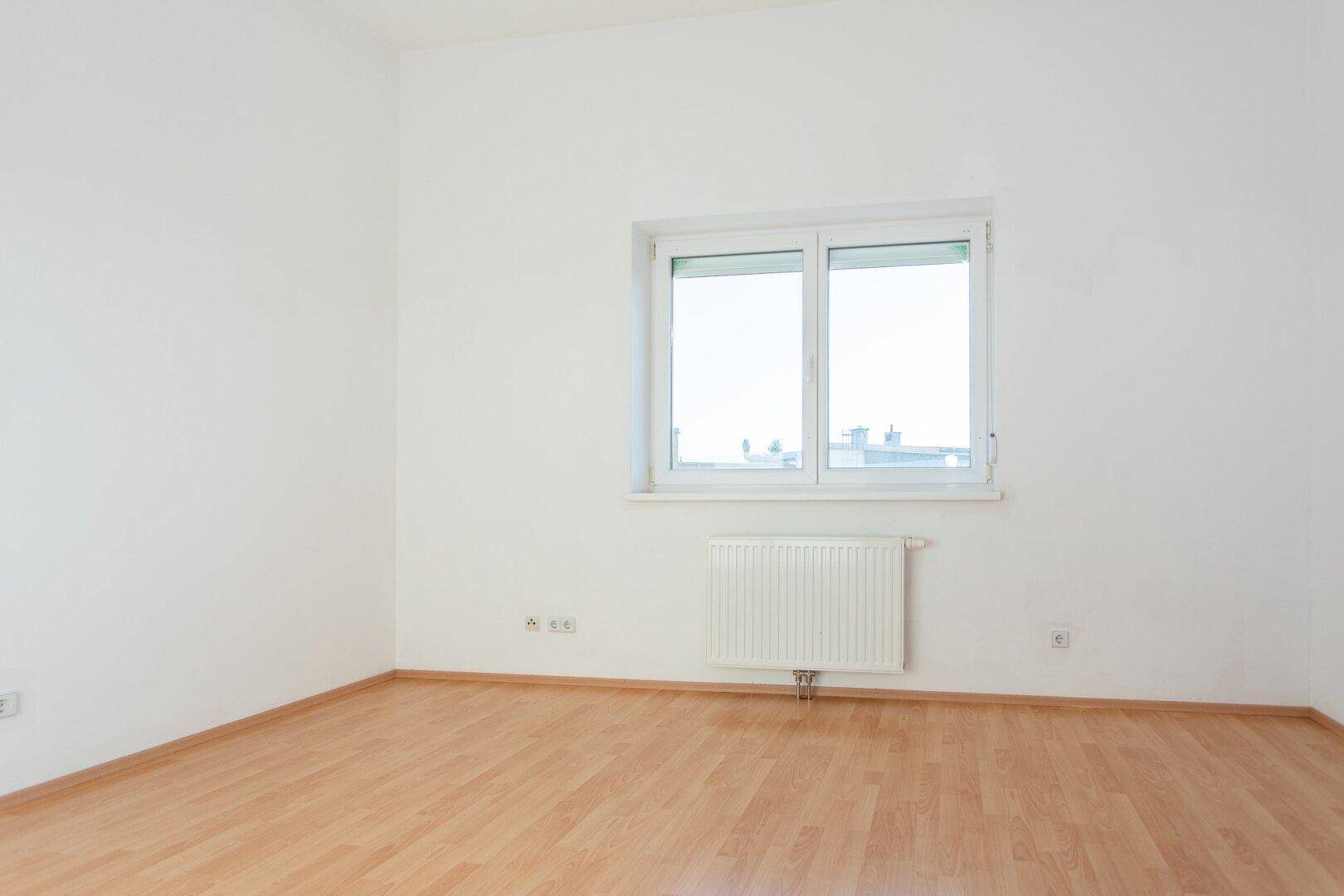 Heller, leerer Raum mit Laminatboden, weiß gestrichenen Wänden und einem Fenster mit Heizkörper.