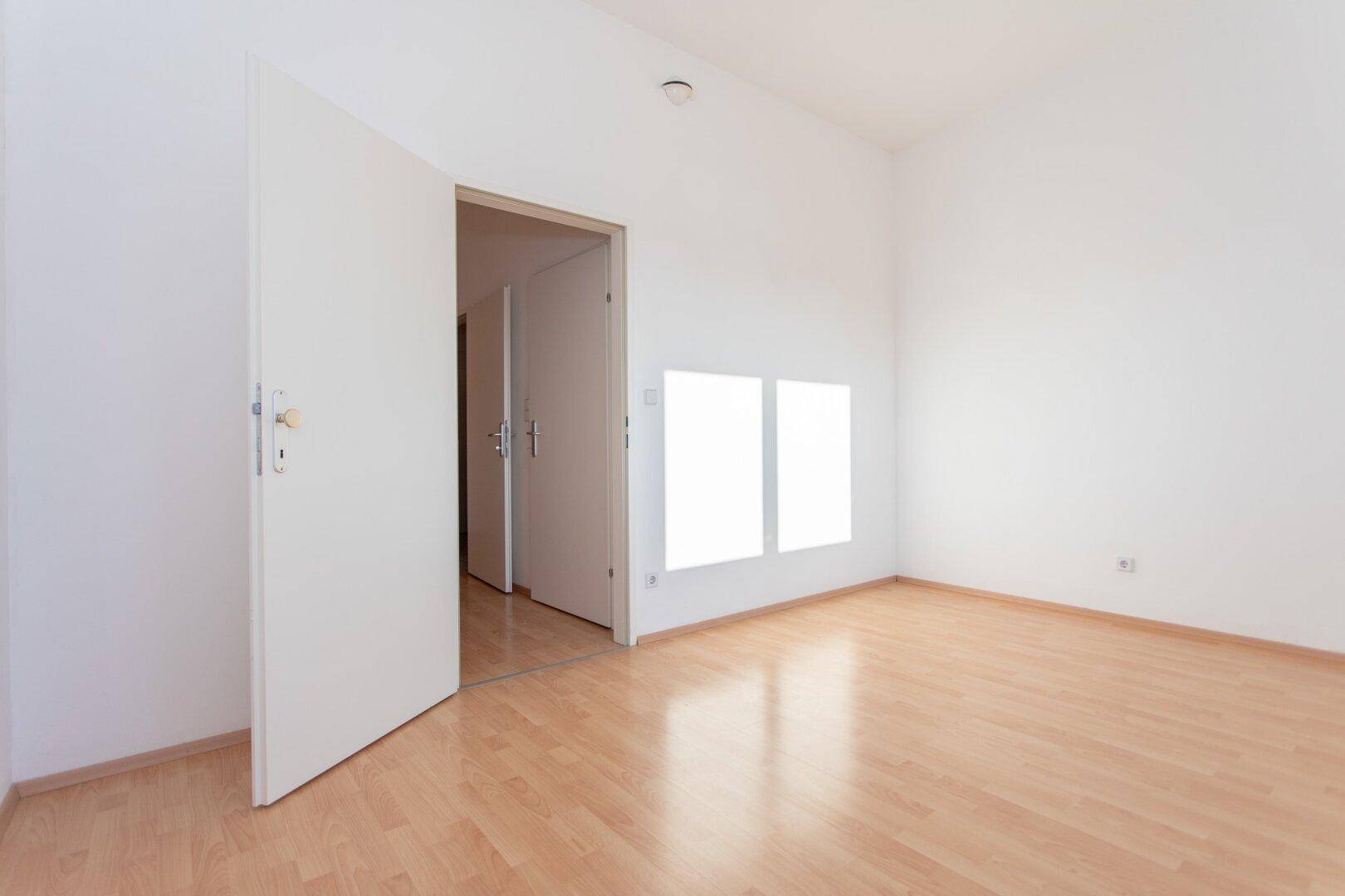 Heller, leerer Raum mit Laminatboden und weiß gestrichenen Wänden, mit offener Tür zum Flur.